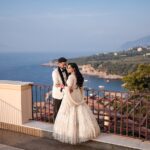 The Bride Wearing A White Dress And The Groom Wearing A White Tuxedo Stand On A Terrace Overlooking The Italian Sorrento Coast Following Their Indian Wedding In Italy